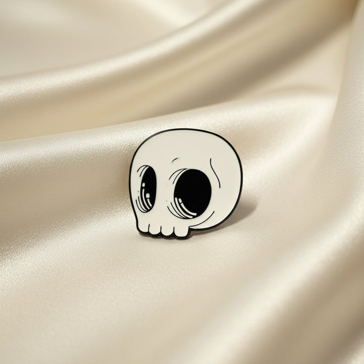 An enamel pin of a stylized cartoon style skull in white and black enamel on a light fabric background.
