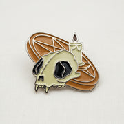 An enamel lapel pin featuring a pentagram altar with a lit candle and a cat skull on top, with an orange pentagram altar, depicted on a white cloth background.