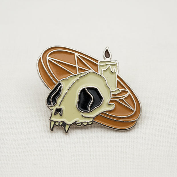 An enamel lapel pin featuring a pentagram altar with a lit candle and a cat skull on top, with an orange pentagram altar, depicted on a white cloth background.