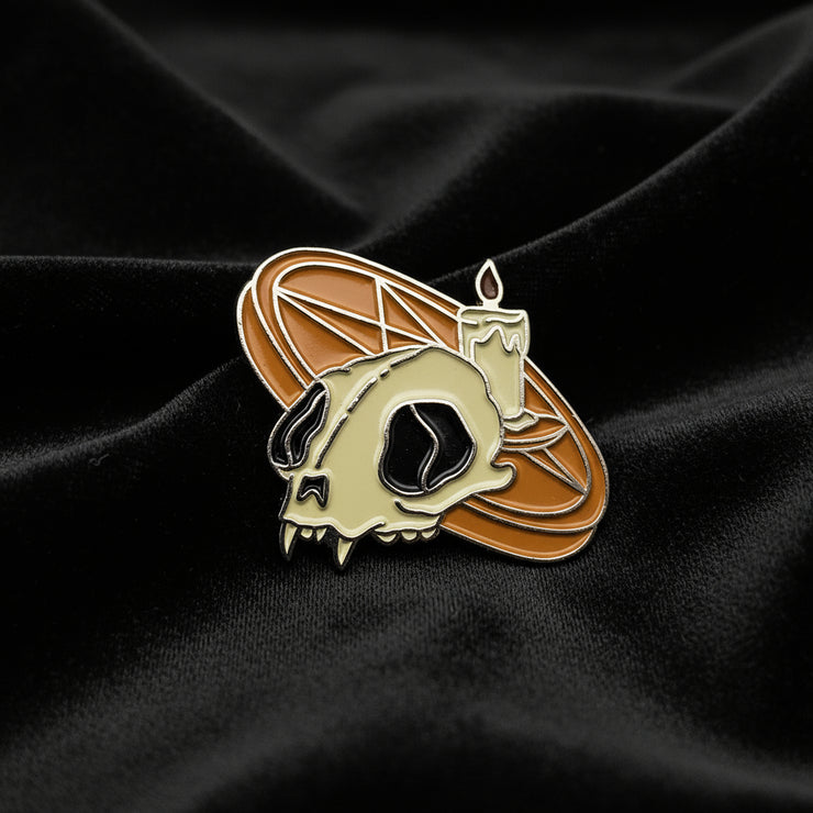 This enamel lapel pin features a vivid orange pentagram altar adorned with a lit candle and a cat skull on a dark fabric background.