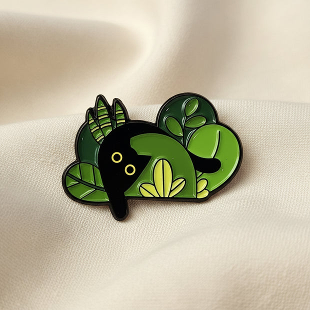 Green and black enamel pin shaped like plants with a black cat hiding in it on a light colored background.