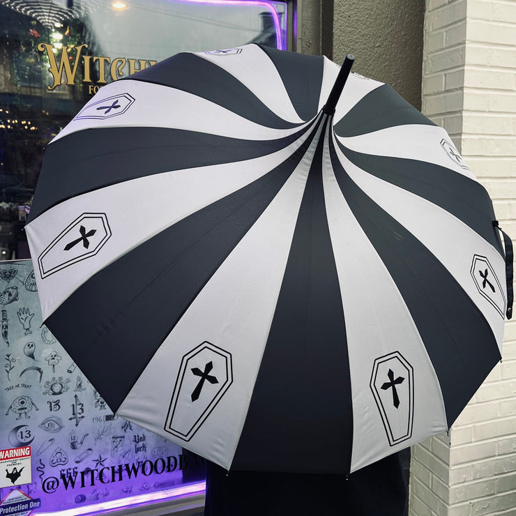Black and white striped umbrella with coffin and cross design, held by a person in front of a store window.