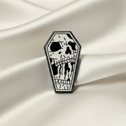 Coffin shaped enamel pin with skull design on a light colored background.