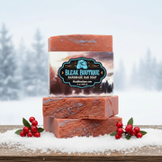 Two bars of Bleak Boutique handmade soap stacked on a snowy surface with berries and a blurred winter background.