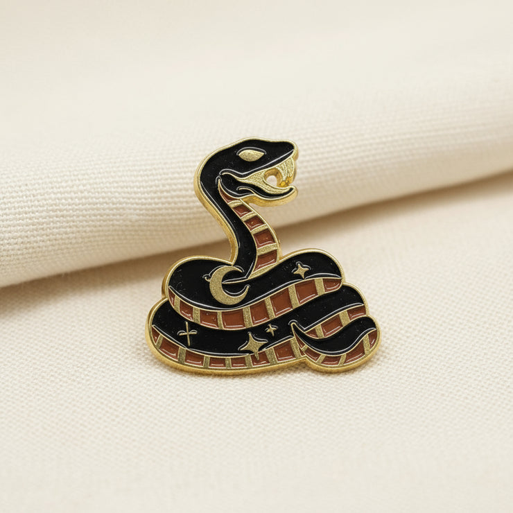 an enamel lapel pin shaped like a snake with a black body, brown scales, and stars on its back is pictured on a light fabric background.