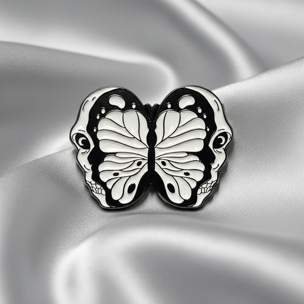 An enamel pin depicting a split open skull revealing a butterfly within on a light colored background