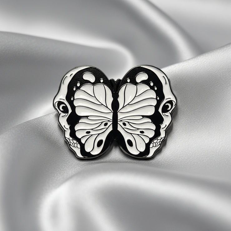 An enamel pin depicting a split open skull revealing a butterfly within on a light colored background