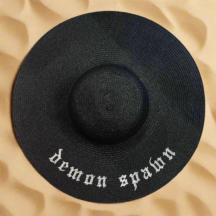 Black straw hat with 'demon spawn' text on a beige sand background
