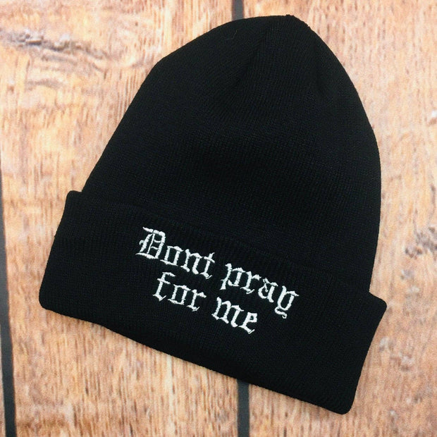 Black beanie with 'Don't pray for me' text on a wooden surface