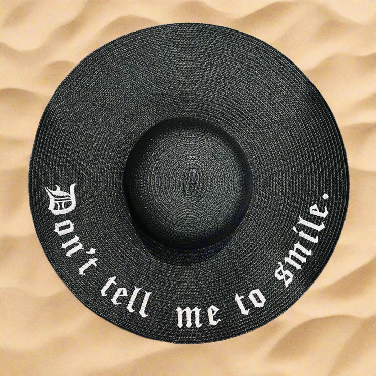 Black straw hat with 'Don't tell me to smile' text on a beach sand background