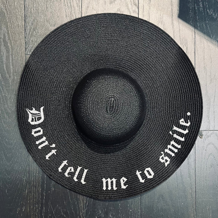 Black straw hat with 'Don't tell me to smile' text on a wooden surface