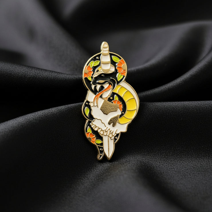 An enamel pin of a snake wrapped around a dagger and skull on a dark background.
