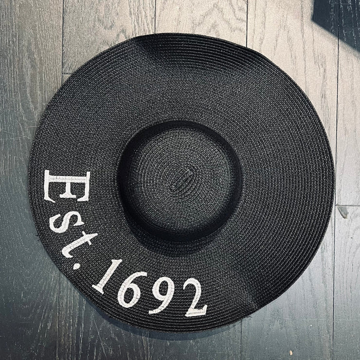 Black straw hat with 'Est. 1692' text on a wooden surface