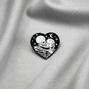 A heart shaped enamel pin depicting a pair of skeletons kissing and embracing under a night sky on a light background.