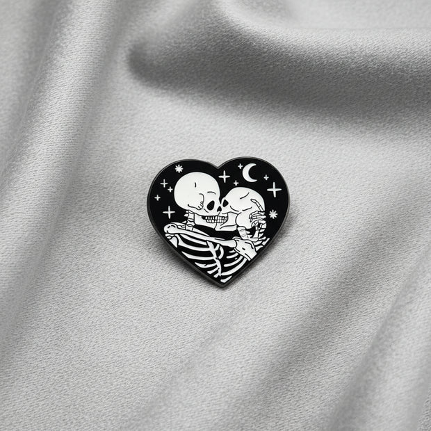 A heart shaped enamel pin depicting a pair of skeletons kissing and embracing under a night sky on a light background.