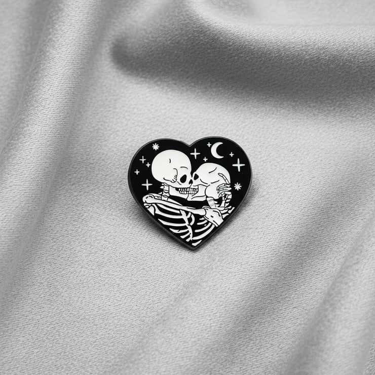 A heart shaped enamel pin depicting a pair of skeletons kissing and embracing under a night sky on a light background.