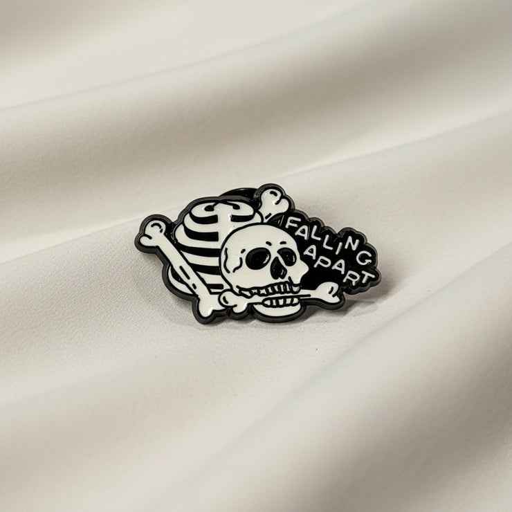 Humorous enamel pin depicting a pile of bones and text reading "Falling Apart" on a light background