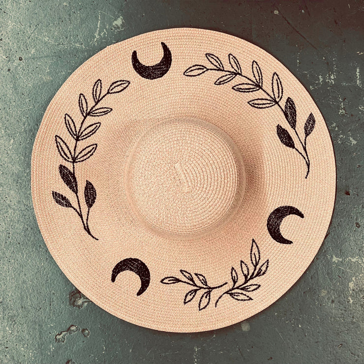 Blush pink straw hat with black leaf and moon designs on a textured surface