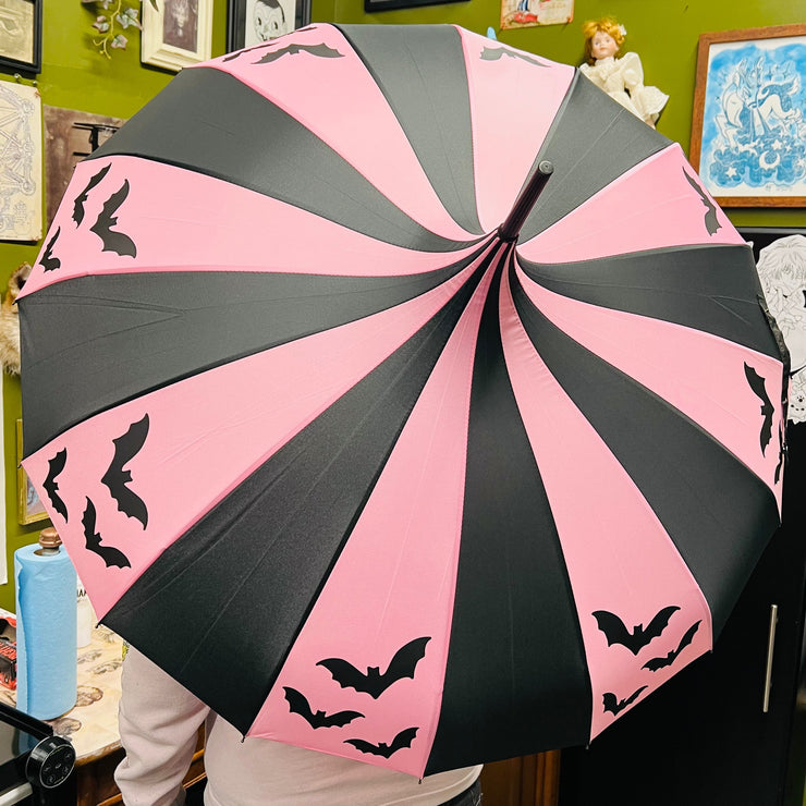 Pink and black striped umbrella with bat designs held by a person in a room with framed pictures on the wall.