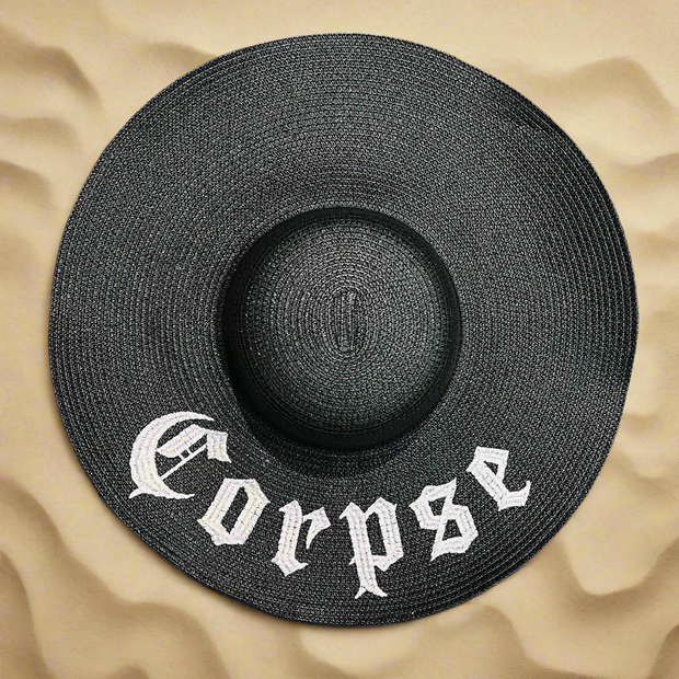 Black straw hat with the word "Corpse" embroidered on it in white thread. The hat rests on a beach sand background.