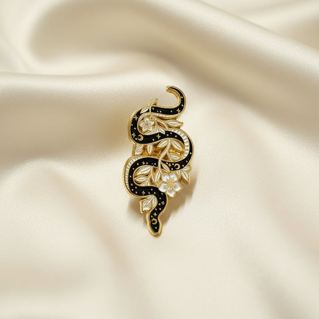 Enamel pin depicting a black snake intertwined with white and gold flowers on a light background