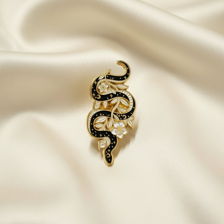 Enamel pin depicting a black snake intertwined with white and gold flowers on a light background