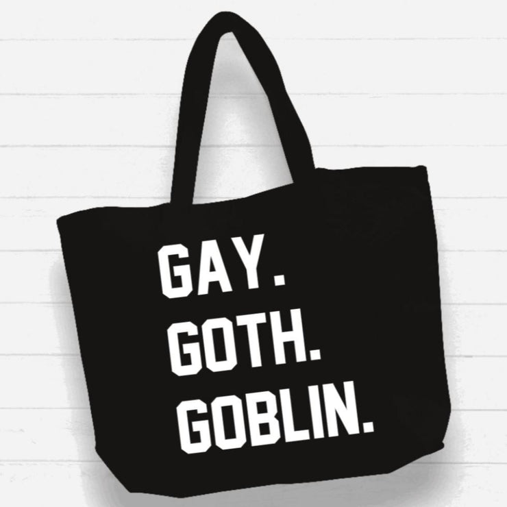 Black tote bag with white text 'GAY. GOTH. GOBLIN.' on a white wooden background