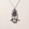 A silver-toned pendant necklace featuring an oval purple stone at the top and two crescent moon pendants below, one of which contains a smaller round purple stone, shown on a light beige background.