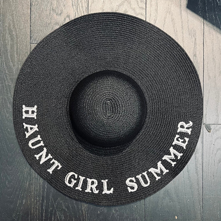 Black straw hat with 'HAUNT GIRL SUMMER' text on a wooden floor.