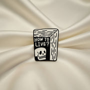 Black and white enamel pin with 'How to Live?' text and skull design on a light gray background