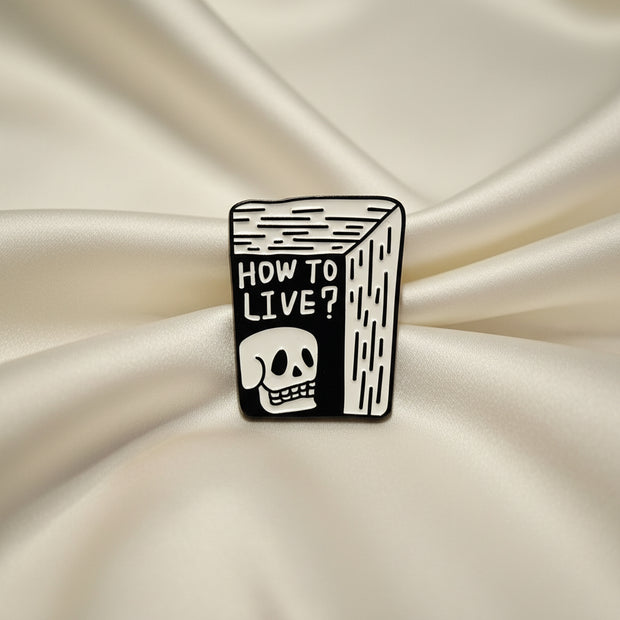 Black and white enamel pin with 'How to Live?' text and skull design on a light gray background