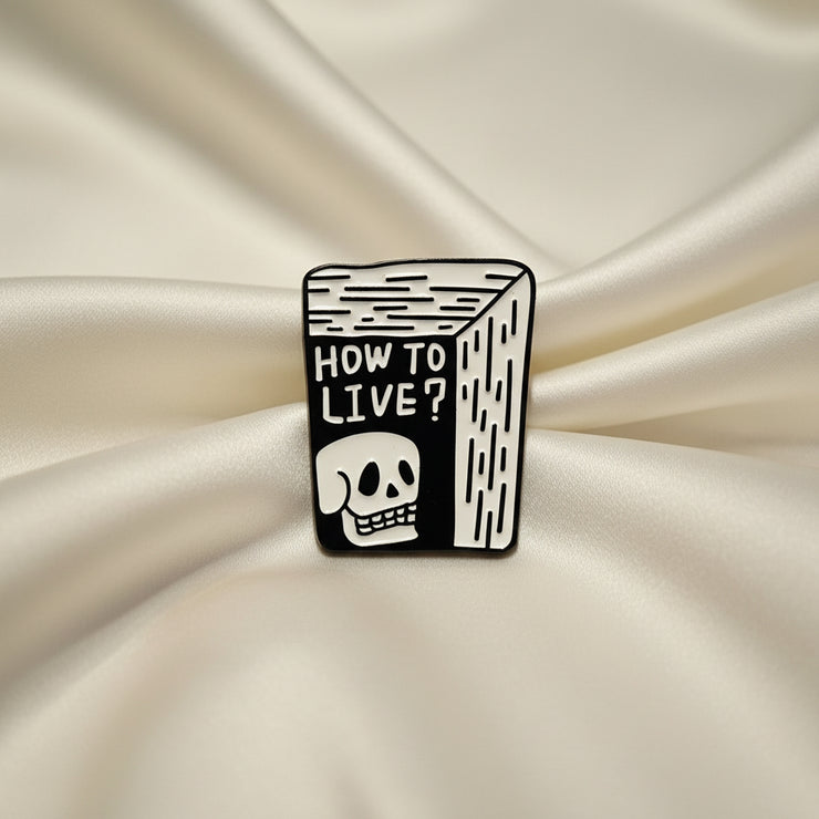Black and white enamel pin with 'How to Live?' text and skull design on a light gray background