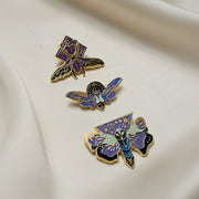 A trio of enamel pins depicting three different types of nocturnal insects: lightning bug, stag beetle, and moth on a light background.