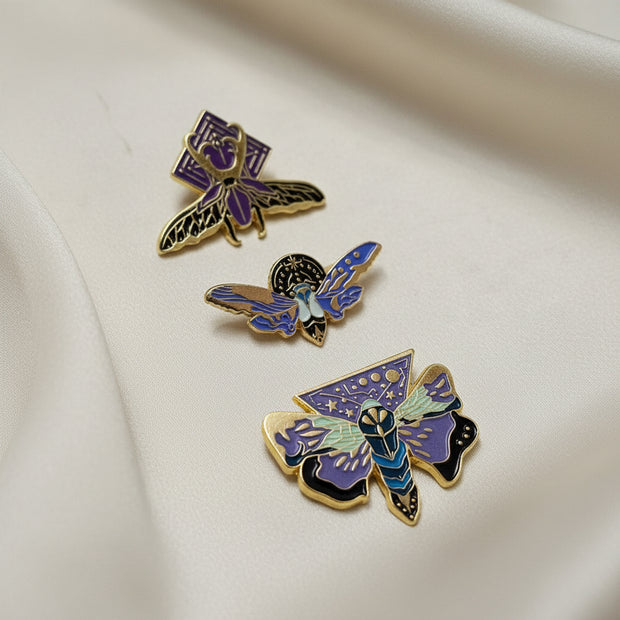 A trio of enamel pins depicting three different types of nocturnal insects: lightning bug, stag beetle, and moth on a light background.
