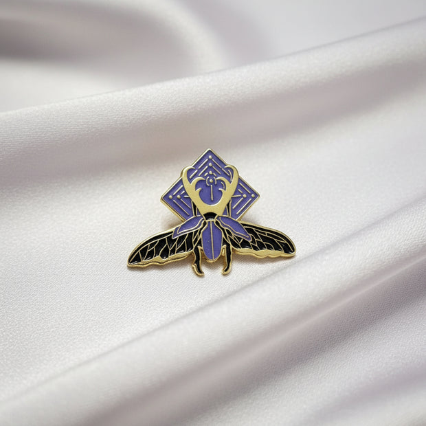 Pin with purple and gold insect design on a beige background