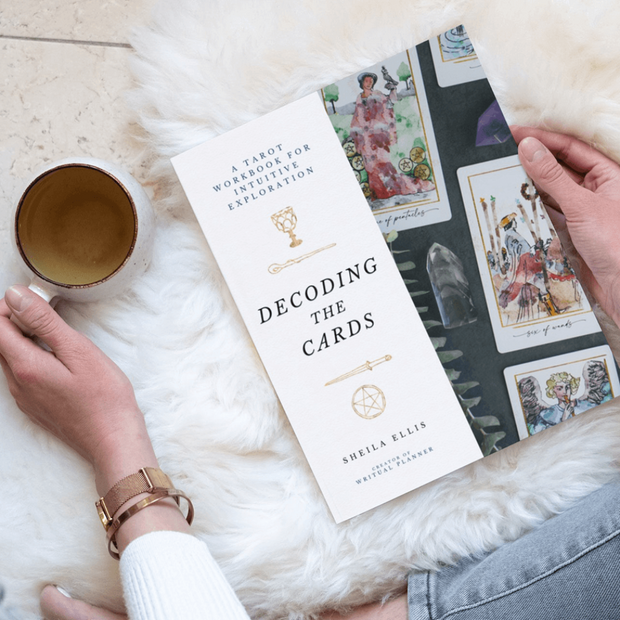 Book titled 'Decoding the Cards' by Sheila Elissa with tarot cards and a cup on a fluffy surface.