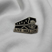 Silver Enamel Lapel Pin depicting a skeleton climbing into a coffin that reads "Life was OK" on a light background.