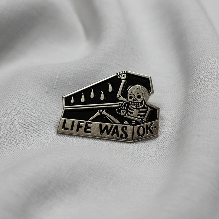 Silver Enamel Lapel Pin depicting a skeleton climbing into a coffin that reads "Life was OK" on a light background.