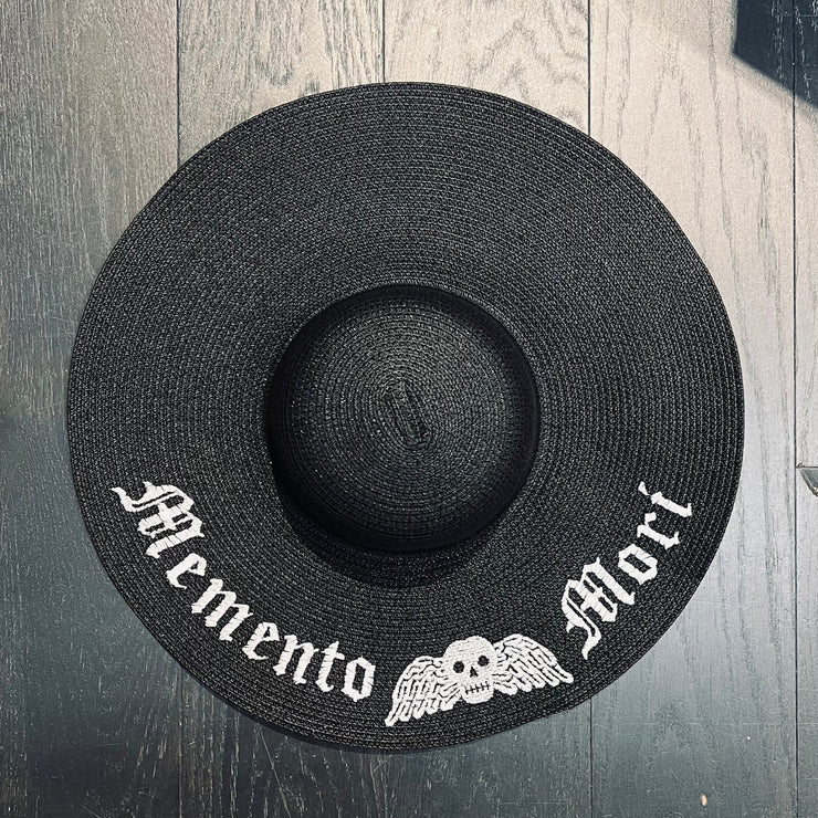 Black straw hat with 'Memento Mori' text and skull design on a wooden surface