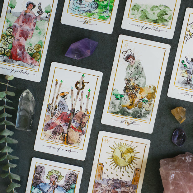 Collection of tarot cards with artistic illustrations on a dark surface with crystals and leaves.