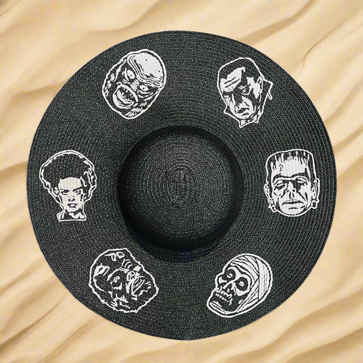 Black straw hat with white embroidered classic horror movie monster faces on a beach sand background