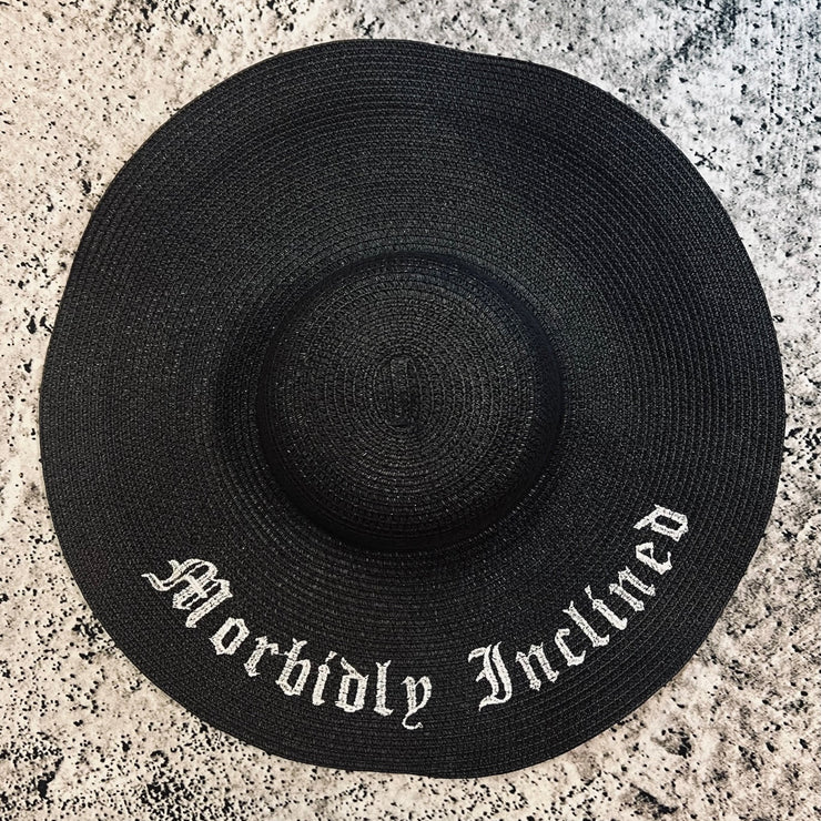 Black straw hat with 'Morbidly Inclined' text on a gray textured surface