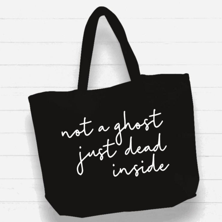 Black tote bag with white text 'not a ghost just dead inside' on a light wooden background