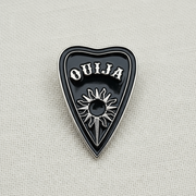The Spirit Board Planchette Enamel Lapel Pin by Bleak Boutique features a black heart-shaped design with OUIJA in white and a sunburst motif, bringing supernatural charm to any outfit.
