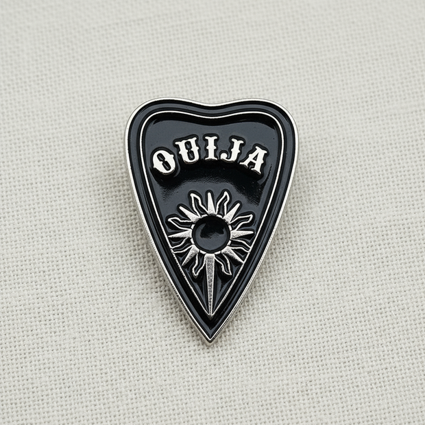 The Spirit Board Planchette Enamel Lapel Pin by Bleak Boutique features a black heart-shaped design with OUIJA in white and a sunburst motif, bringing supernatural charm to any outfit.