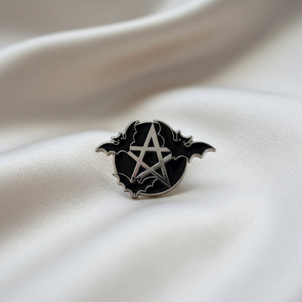 Enamel lapel pin depicting a silver pentacle on a black background with three bats flying around it on a light background.
