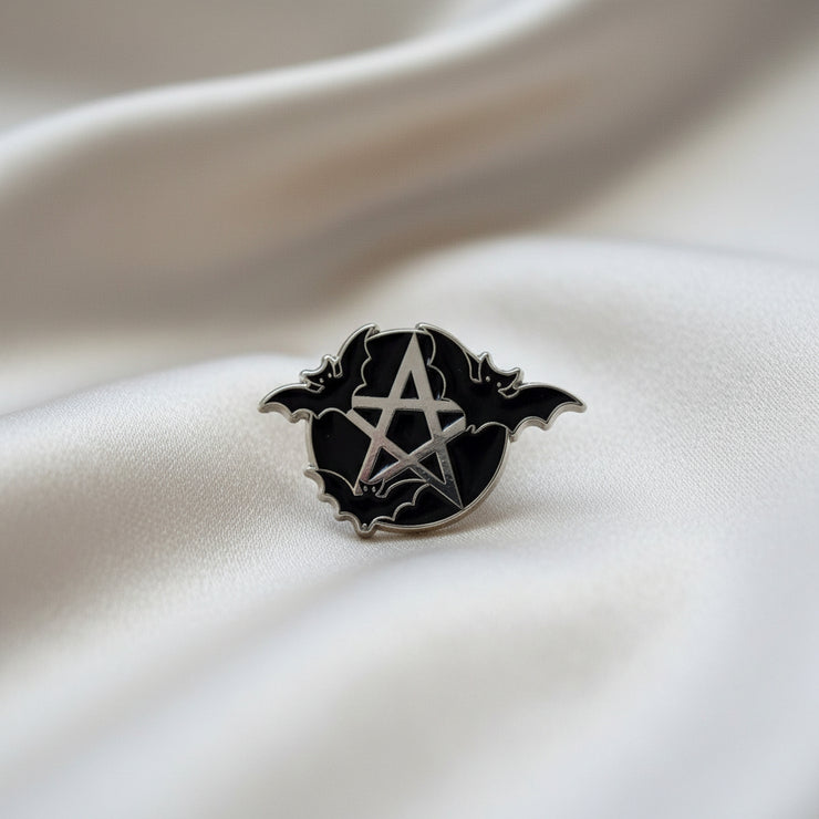 Enamel lapel pin depicting a silver pentacle on a black background with three bats flying around it on a light background.