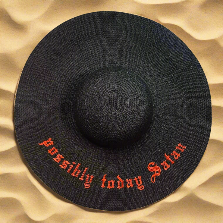 Black straw hat with "Possibly Today Satan" in red text on a beach sand background