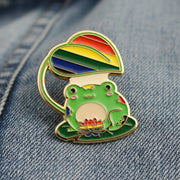 An enamel pin with a green cartoon frog surrounded by rainbow flowers and leaves on a denim background.