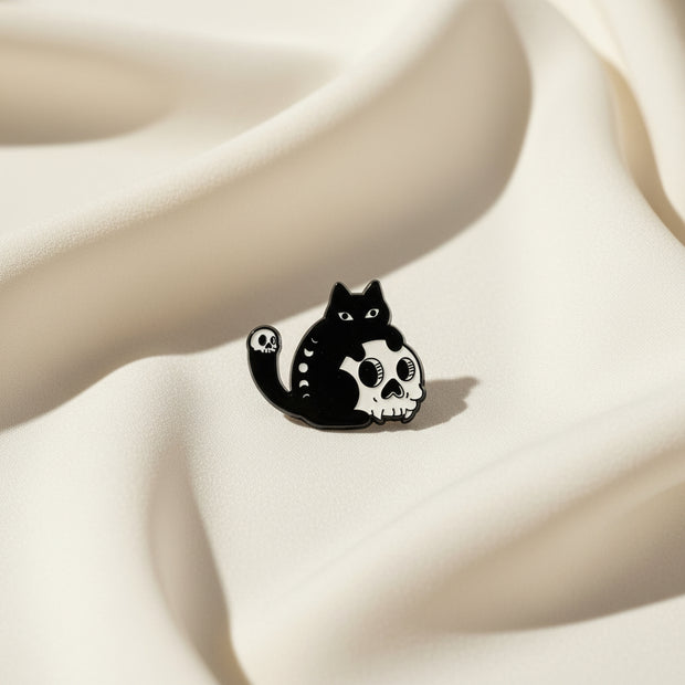 Enamel pin depicting a black cat holding a human skull in its paws on a light background